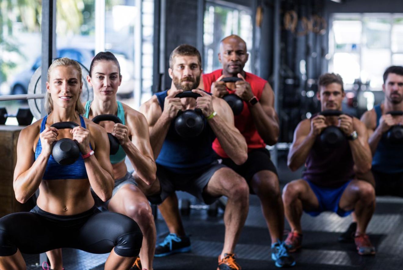 Here is a photo of athletes lifting kettlebells at the gym. 
                        There are two ladies and three men with black kettlebells and colourful outfits.
                        